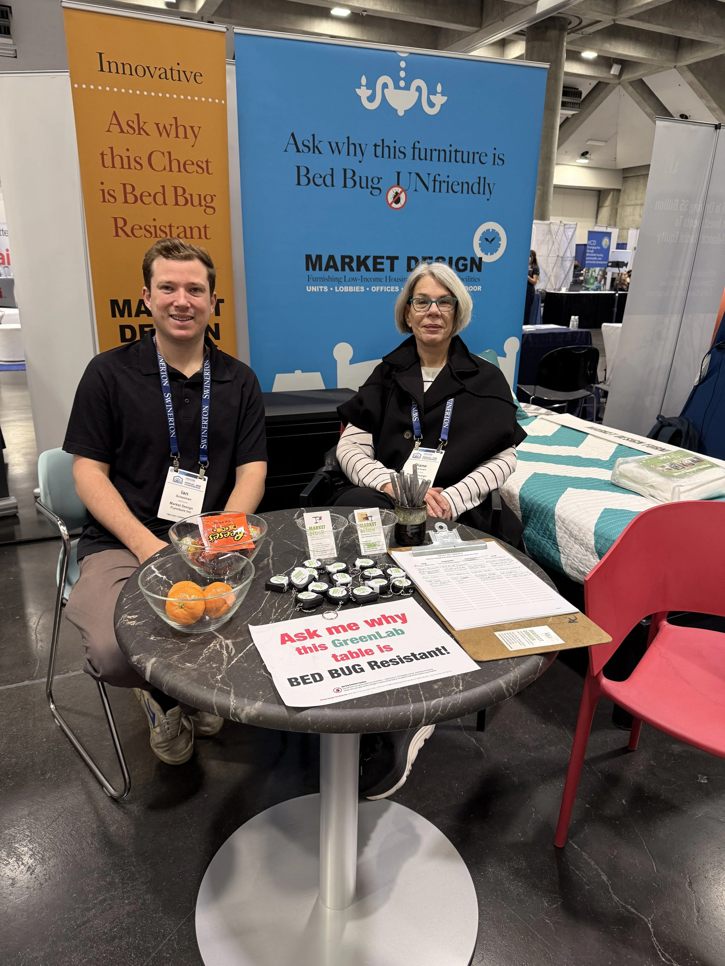 Two people sitting at a booth with a sign that reads ask why this Greenlab table is BED BUG Resistant. There are bowls of oranges, small containers, and papers on the table. The backdrop displays a blue banner with a chandelier and text about furniture being Bed Bug UNfriendly.