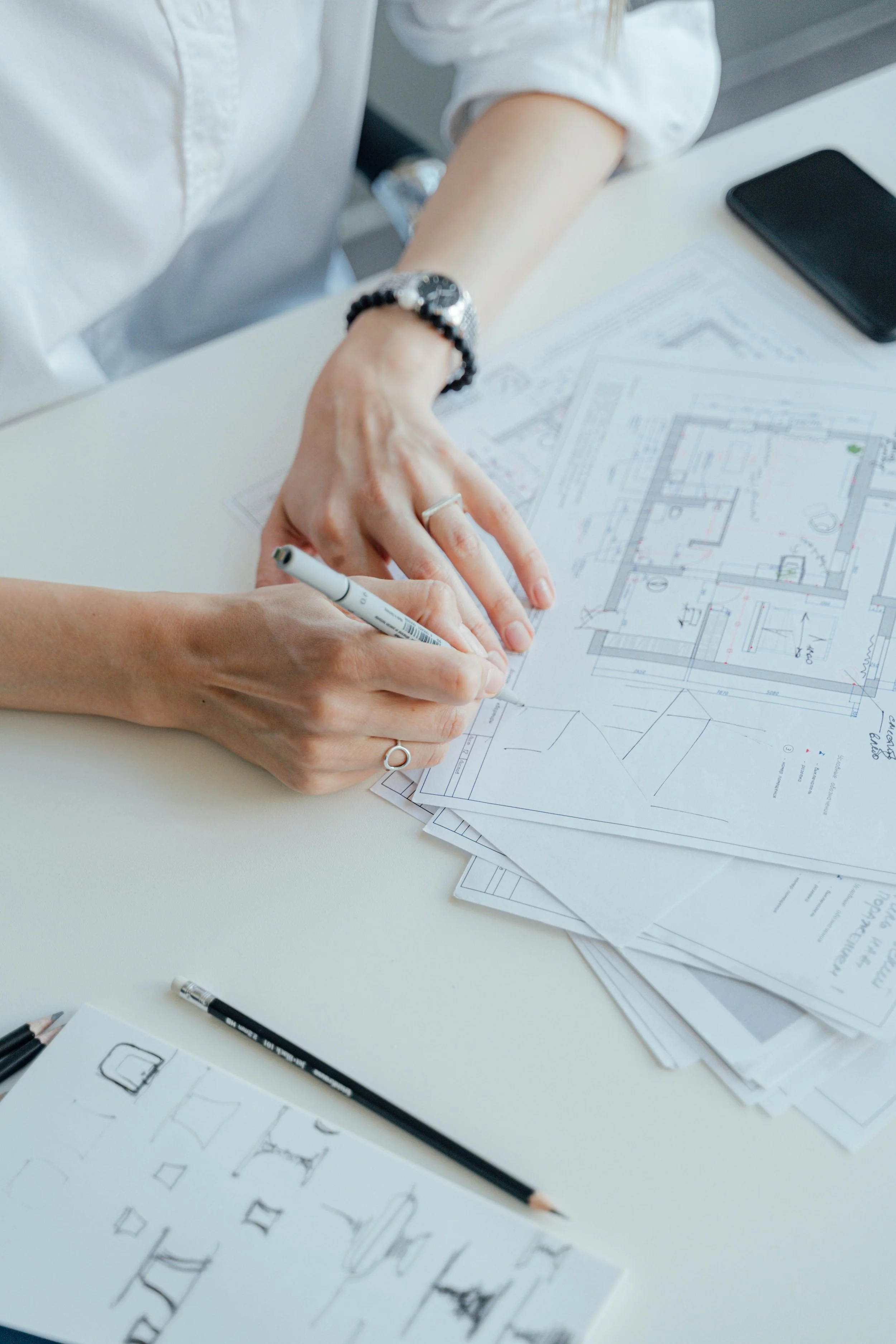 Person reviewing and sketching architectural blueprints on a desk, with sketches and pencils nearby.
