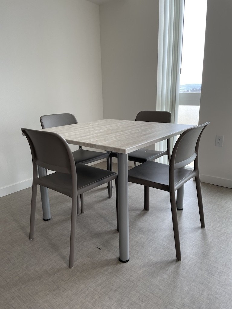 A small dining area with a rectangular table and four gray chairs in a room with white walls, beige carpet, and a window with vertical blinds.