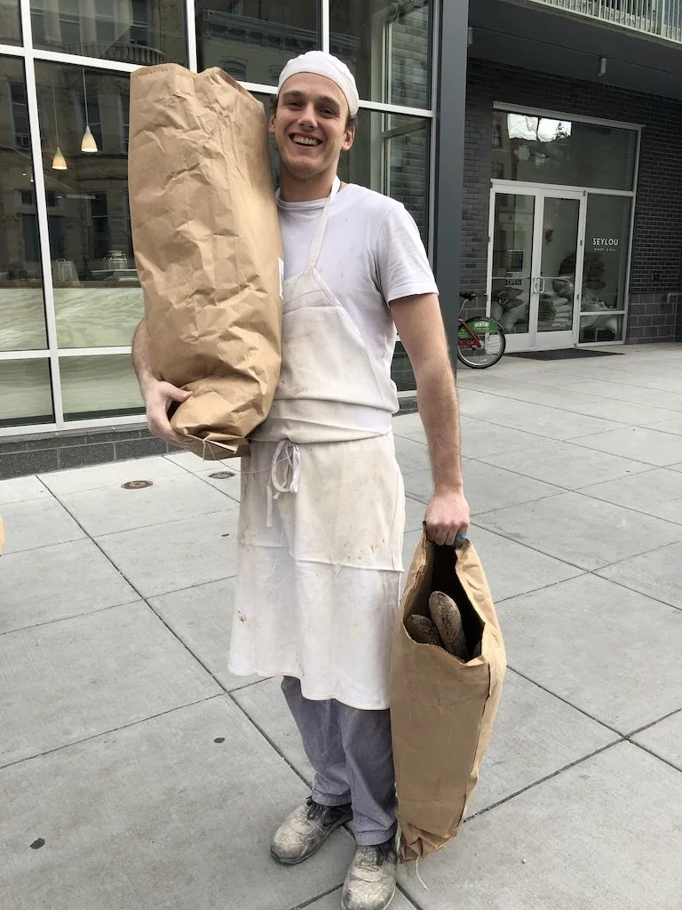 Photo of a smiling bakery employee holding a donation of bread and baguettes for DC Food Rescue