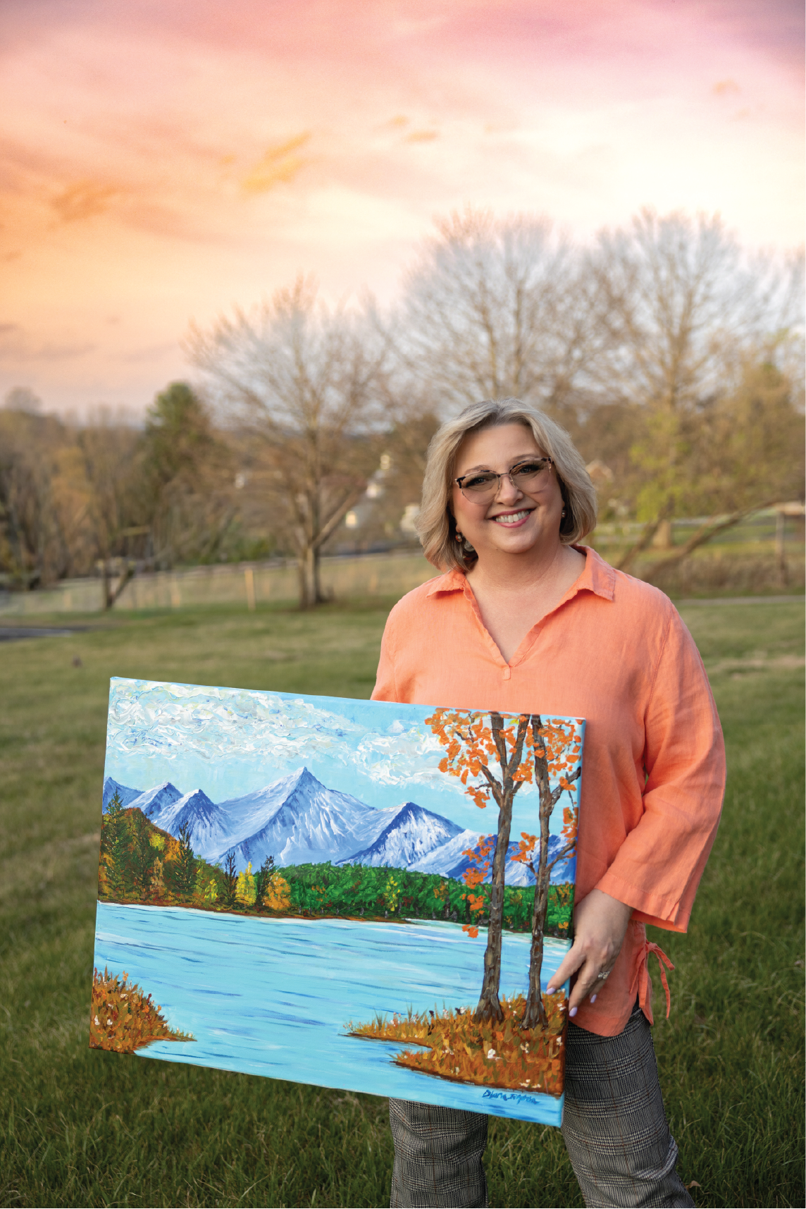 A woman holding a colorful landscape painting of mountains, trees, and water outdoors during sunset.