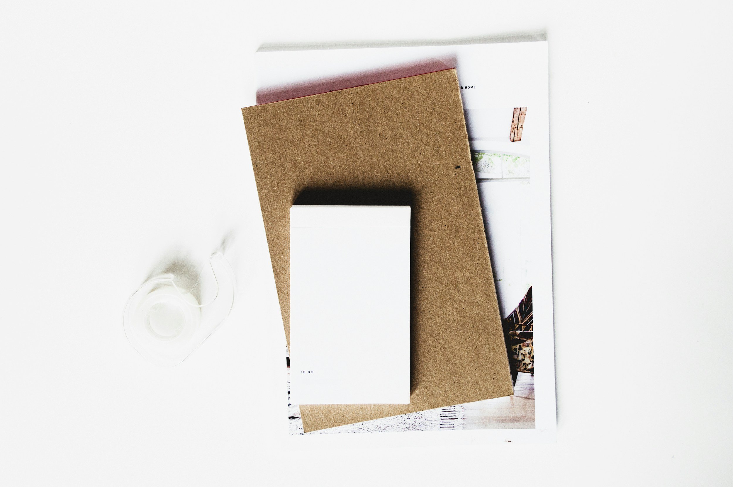 Top view of a desk with various stationery and magazine, featuring a cork notebook, a white paper, and a roll of transparent tape on a white surface.