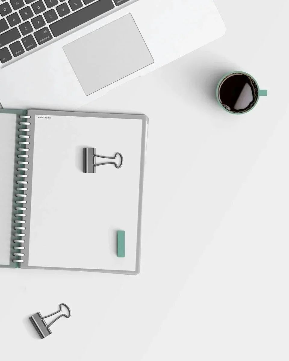 Top-down view of a workspace with a laptop, a coffee mug, a spiral notebook, and binder clips on a white surface.