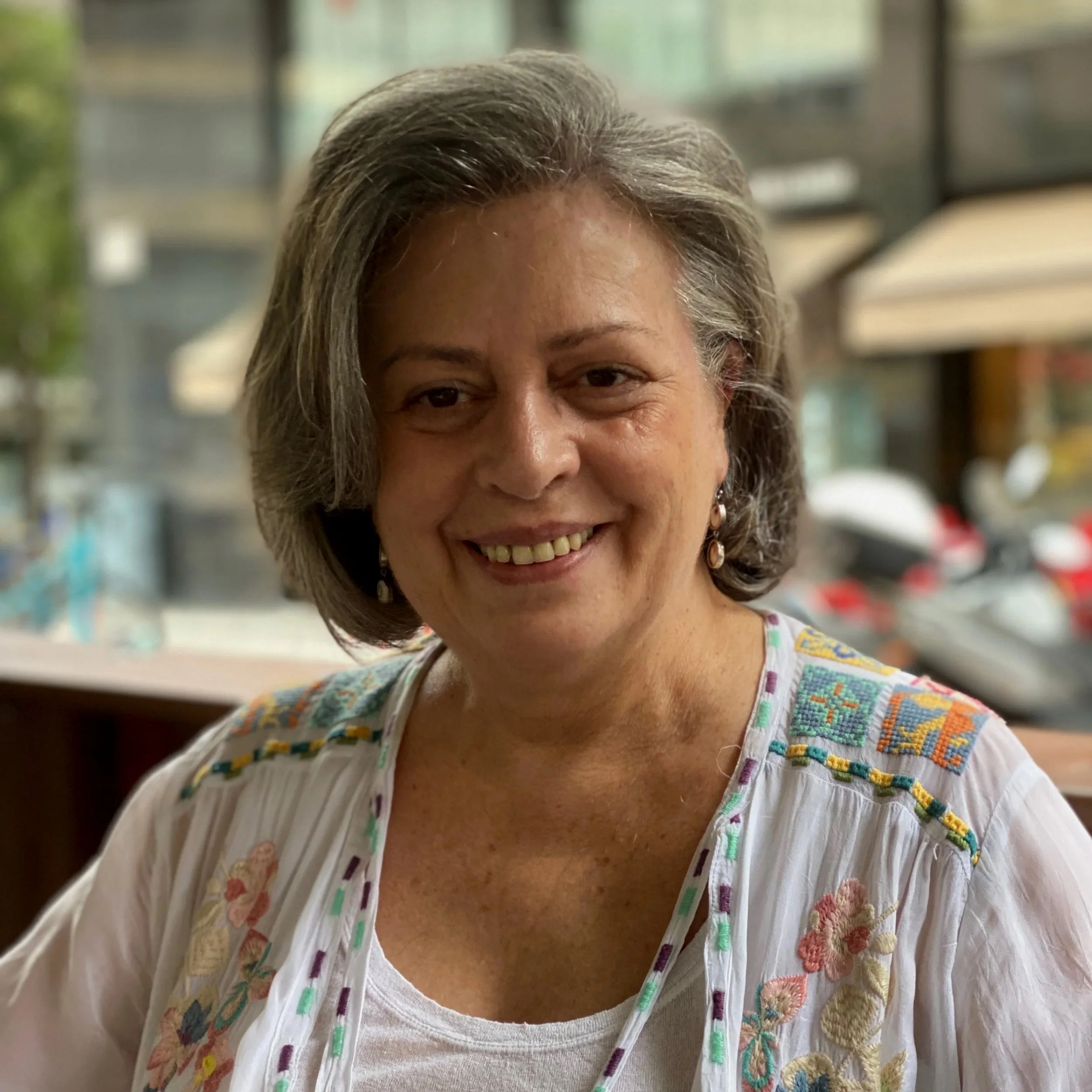 A smiling woman with gray hair, wearing a white embroidered blouse, sitting indoors near a window with a street scene visible outside.