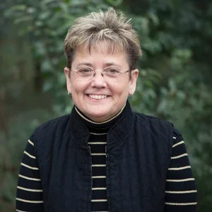 Smiling woman outdoors with trees in background, wearing glasses, a black vest, and striped shirt.