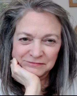 Close-up of a woman with gray hair resting her chin on her hand, looking at the camera.