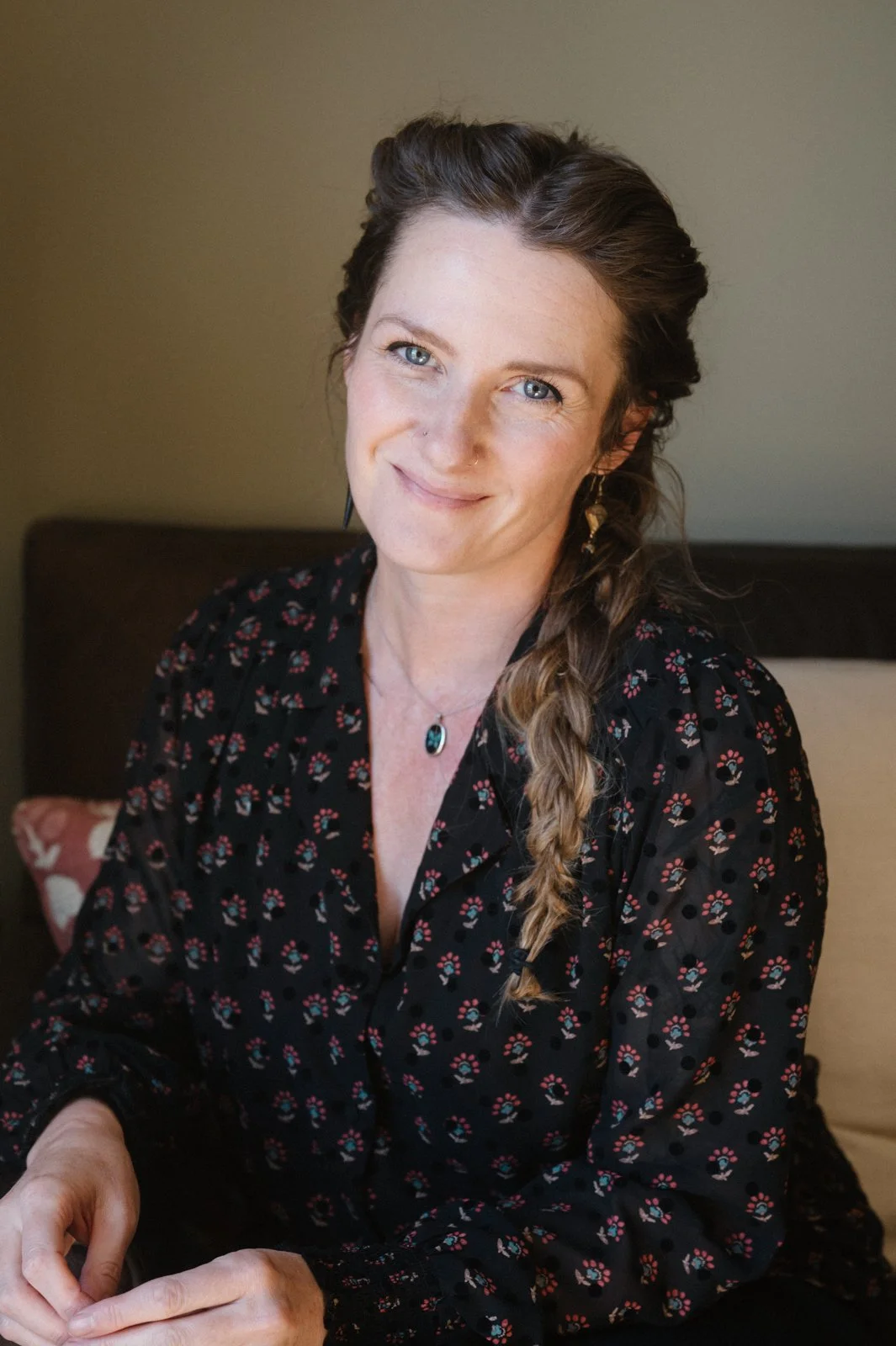 A woman with long, braided brown hair, wearing a black dress with a colorful floral pattern, gold earrings, a black necklace with a blue gemstone, and sitting on a bed with a beige and pink pillow behind her.
