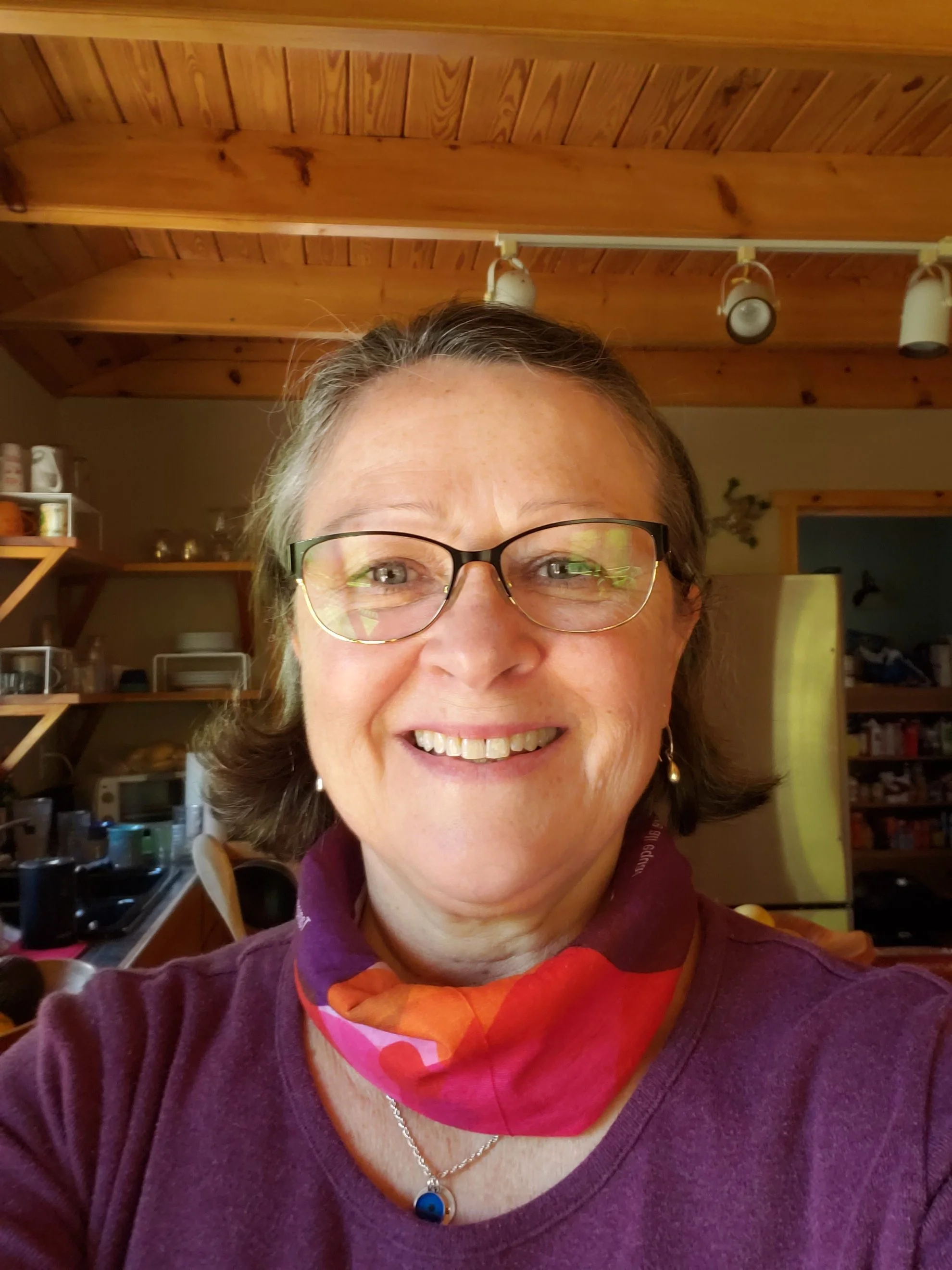 A woman with glasses and short gray hair smiling in a kitchen with wooden ceiling and shelves. She is wearing a colorful scarf, a purple shirt, earrings, and a necklace.