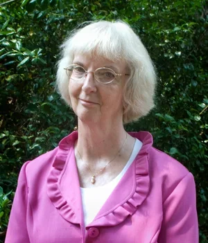 A woman with white hair and glasses standing outdoors with green foliage in the background, wearing a pink shirt.