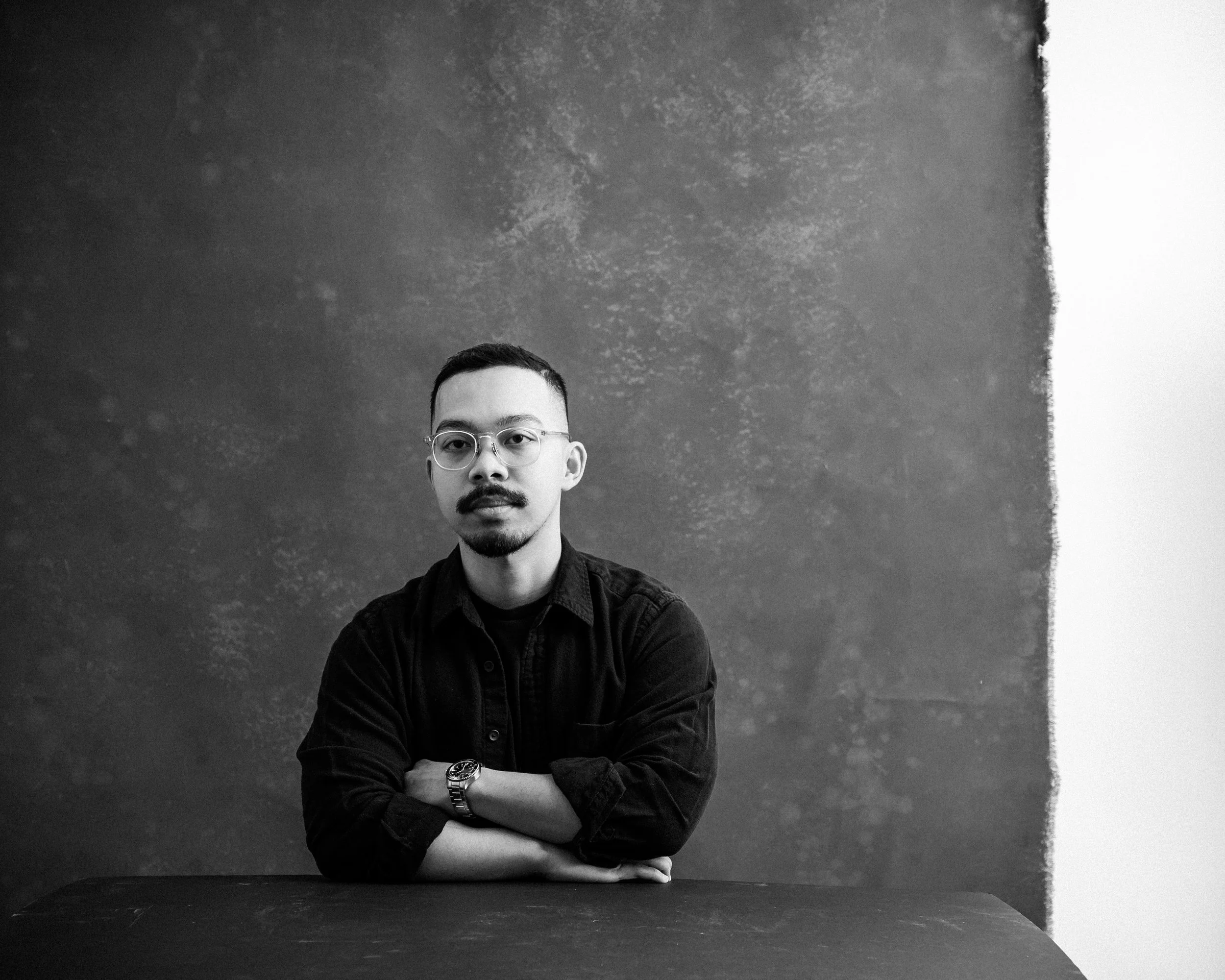John sits at a table facing the camera, hands crossed in front of him in a relaxed pose.