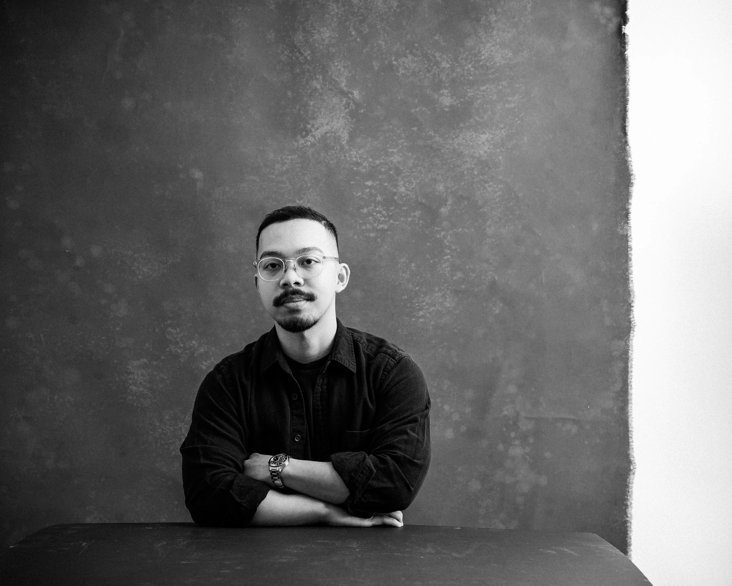 John sits at a table facing the camera, hands crossed, in a composed pose.