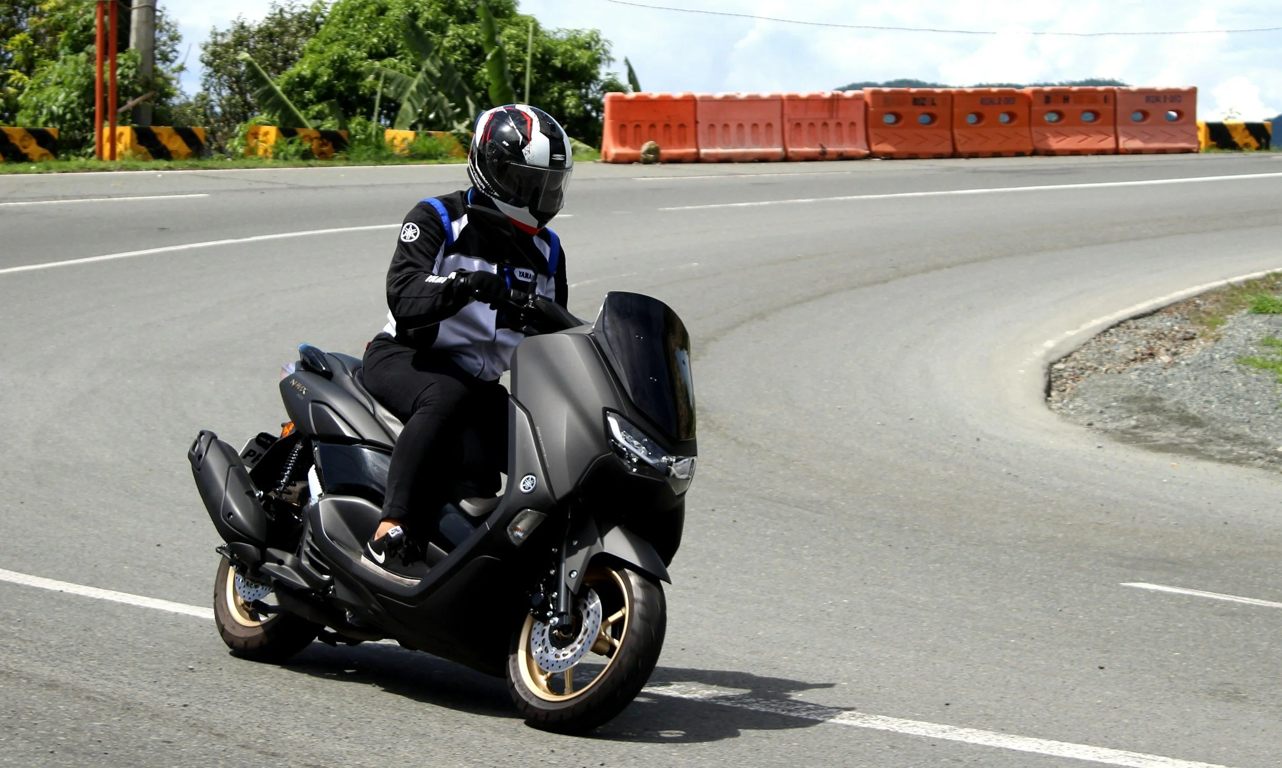 Person riding a black Yamaha scooter on a curved road, wearing black riding gear and a black helmet with white and red accents, with orange safety barriers and green foliage in the background.