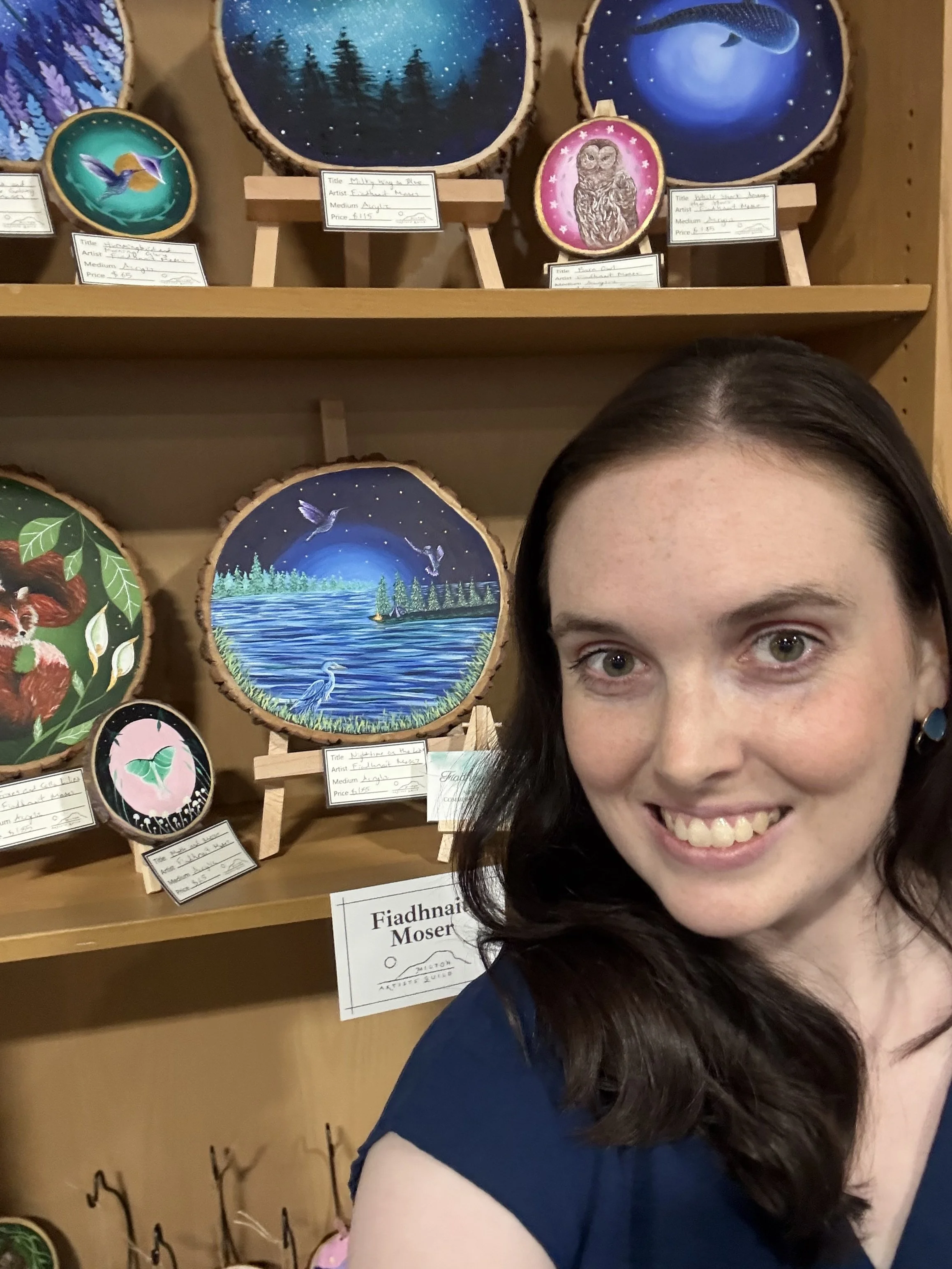 Colorful painted wood art pieces of nature scenes and animals displayed on shelves in an art gallery, with a smiling artist.
