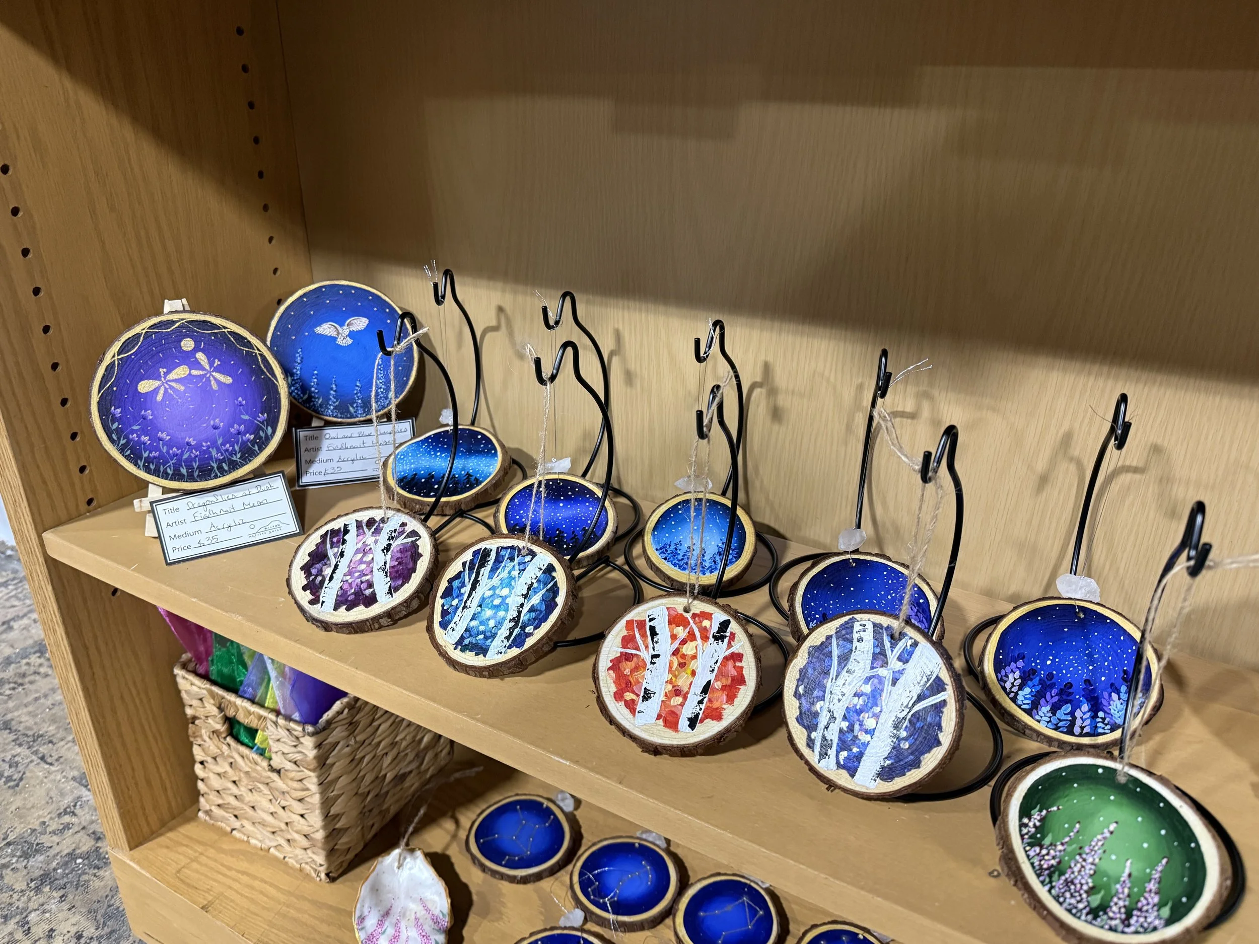 Colorfully painted decorative wooden ornaments with painted scenes of nature, trees, and starry patterns, displayed on a wooden shelf with hooks.