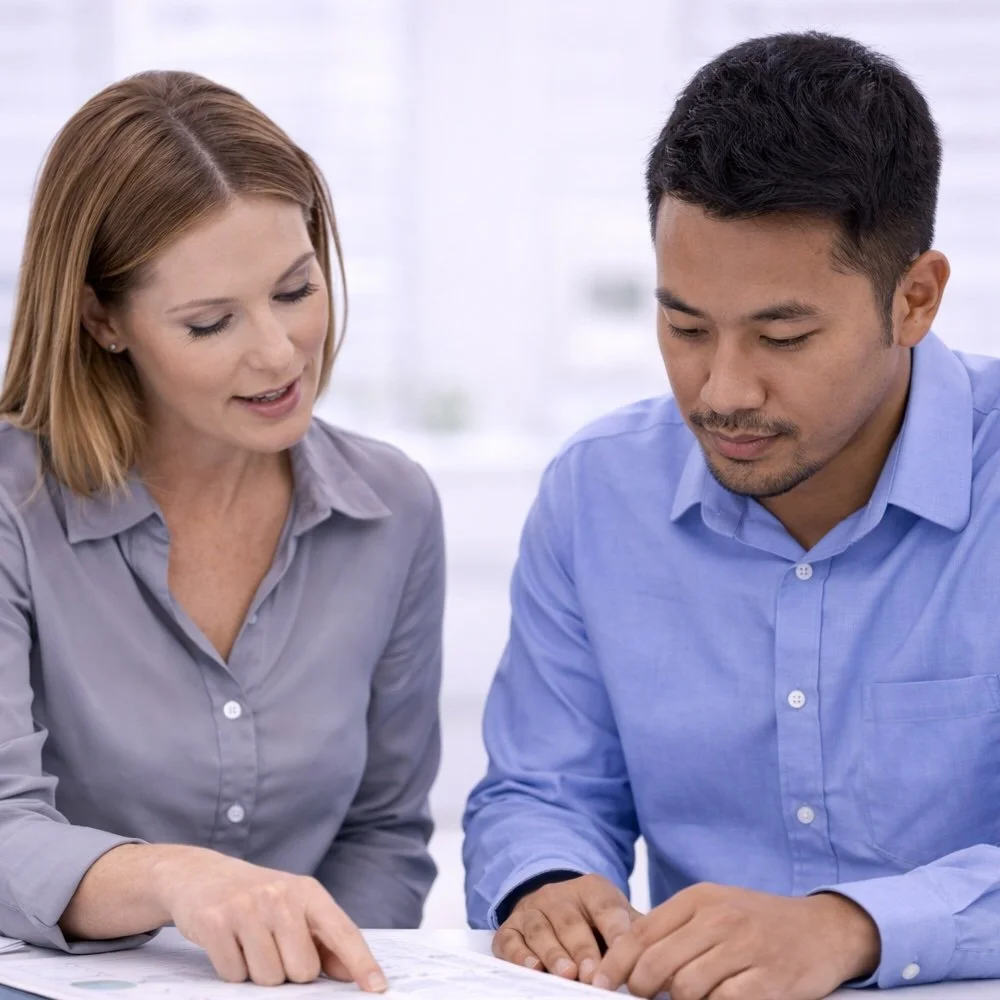 A woman, who is a sales manager, and a man, who is a sales representative, are sitting together, looking at documents or a book on a table, engaged in a discussion or explanation.