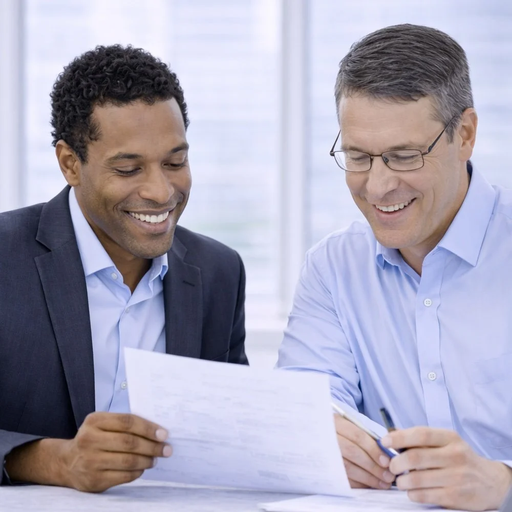 Two men, one who is a sales consultant and one who is a customer, in business suits smiling and reviewing documents together in an office setting.