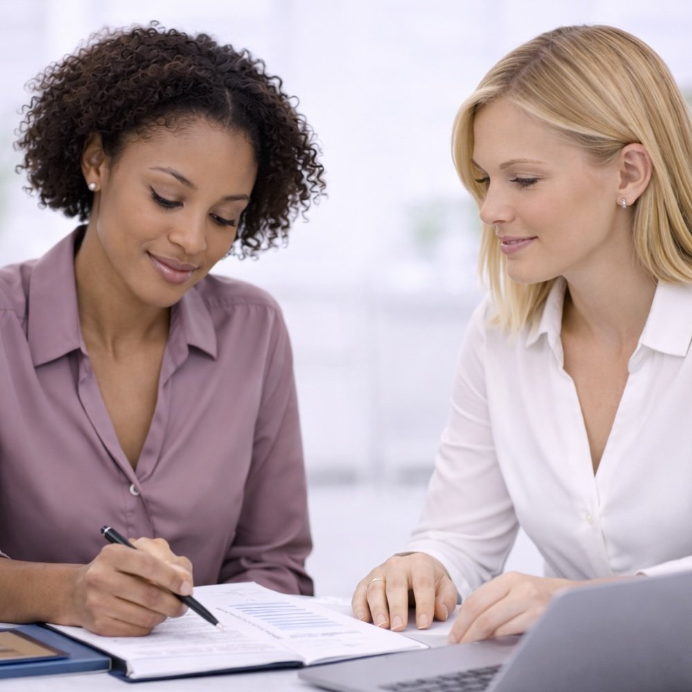 Two sales professionals, both women sitting at a table, reviewing documents together; one is writing, and the other is looking on in an office setting.