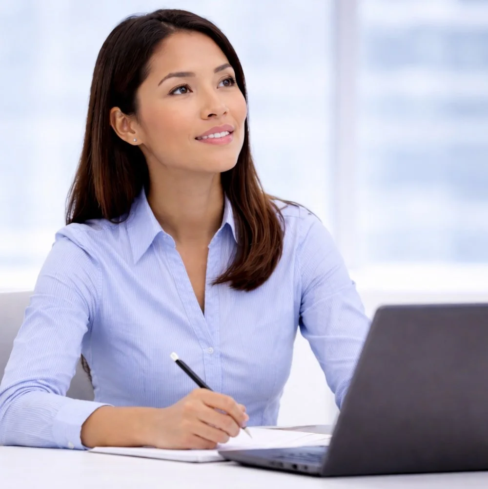A sales professional, woman with brown hair and a blue shirt is sitting at a desk with a laptop and a notepad, holding a pen, looking thoughtfully to her left.
