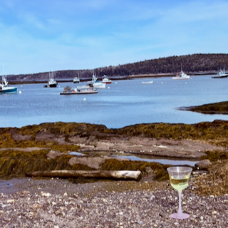 A Coastal Maine waterfront scene with several sailboats anchored on a calm ocean. The foreground features a rocky shoreline with a glass of Chardonnay wine, and the background includes a forested shoreline and a partly cloudy sky.