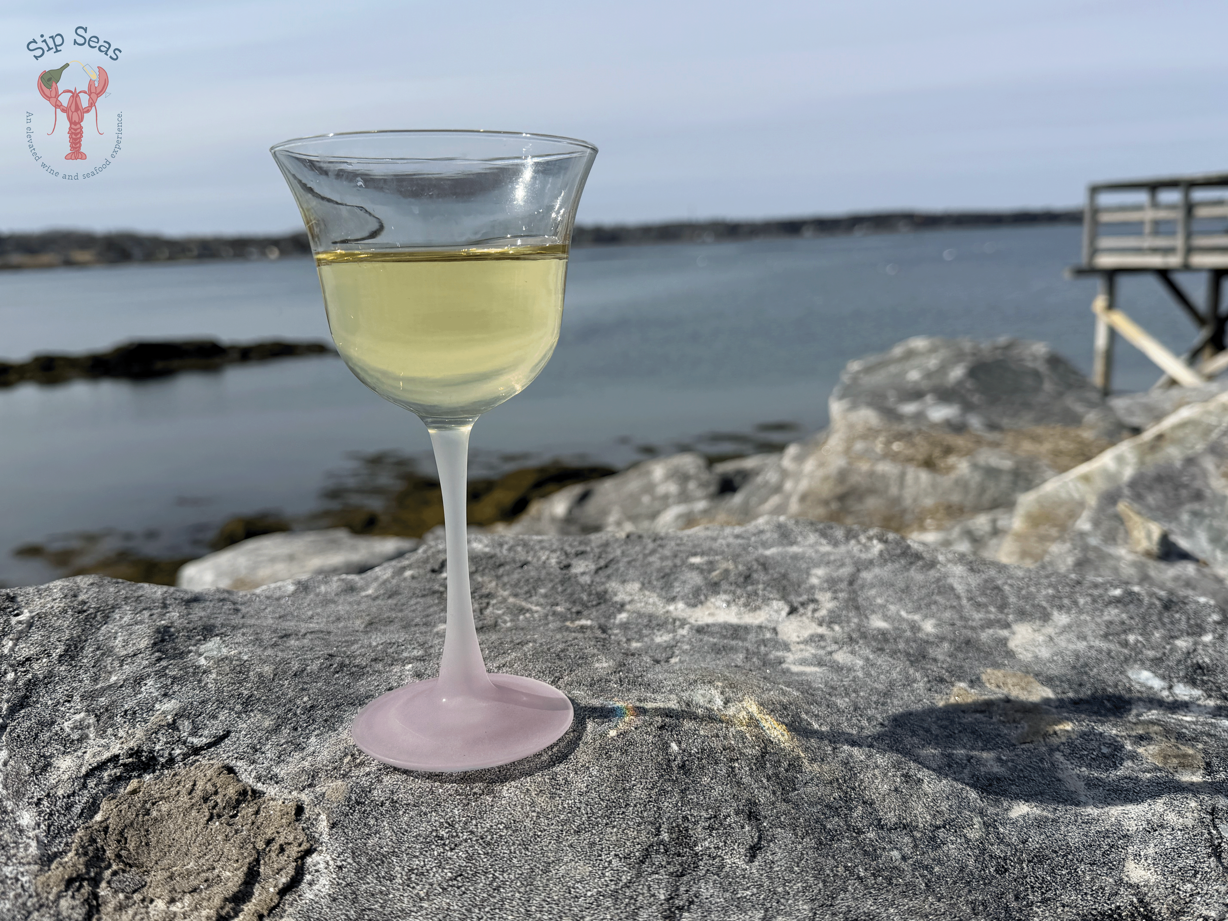 A glass of white Chardonnay wine on a large rock by the water on the Maine coast with a wooden pier or dock in the background and the Sip Seas logo in the top left corner.