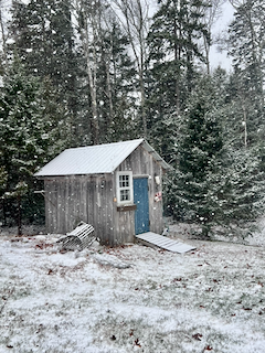 A small wooden shed in a snowy Maine yard surrounded by pine trees.
