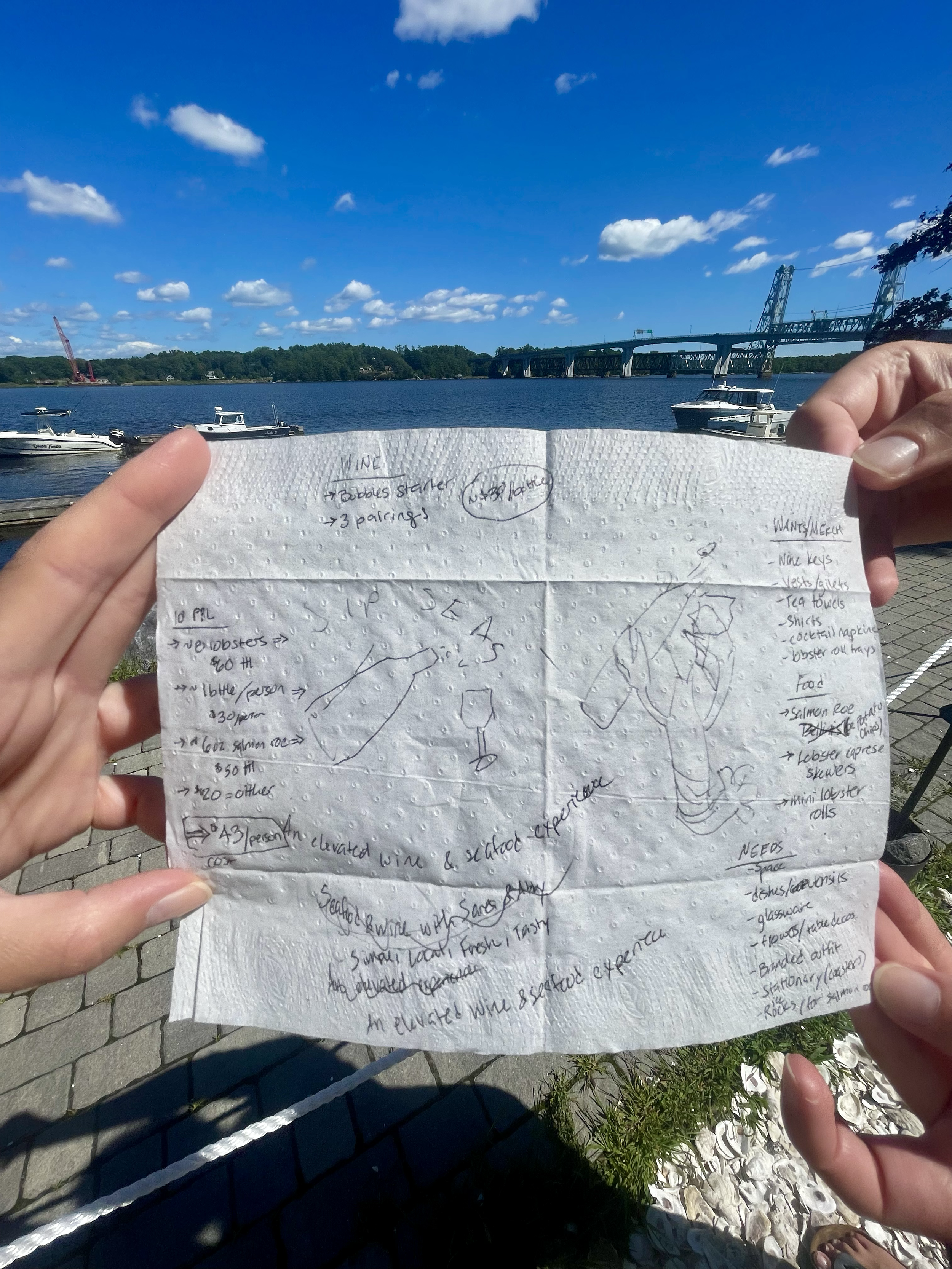 The original idea napkin!  Two female co-founders holding a handwritten note on a napkin outdoors on the Maine coast with the water, boats, a bridge, and trees under a vivid blue sky with scattered clouds in the background.