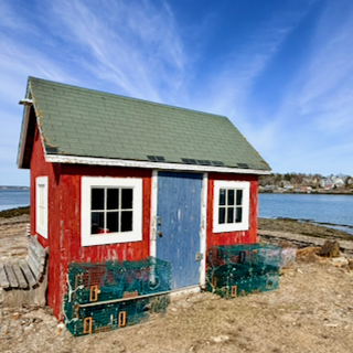 Small red and blue shed with white window frames, beside the water, under a blue sky with clouds on the coast of Maine. There is a bench and stacked lobster traps outside the shack.