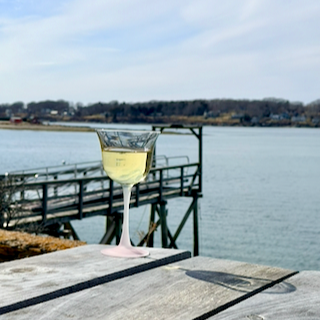 A glass of Chardonnay wine on a wooden picnic table overlooking the Maine coast with a dock, the ocean, and trees in the distance.