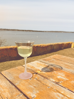 A glass of Chardonnay wine on a weathered wooden picnic table outdoors with the Maine coast, water, and a distant shoreline in the background.
