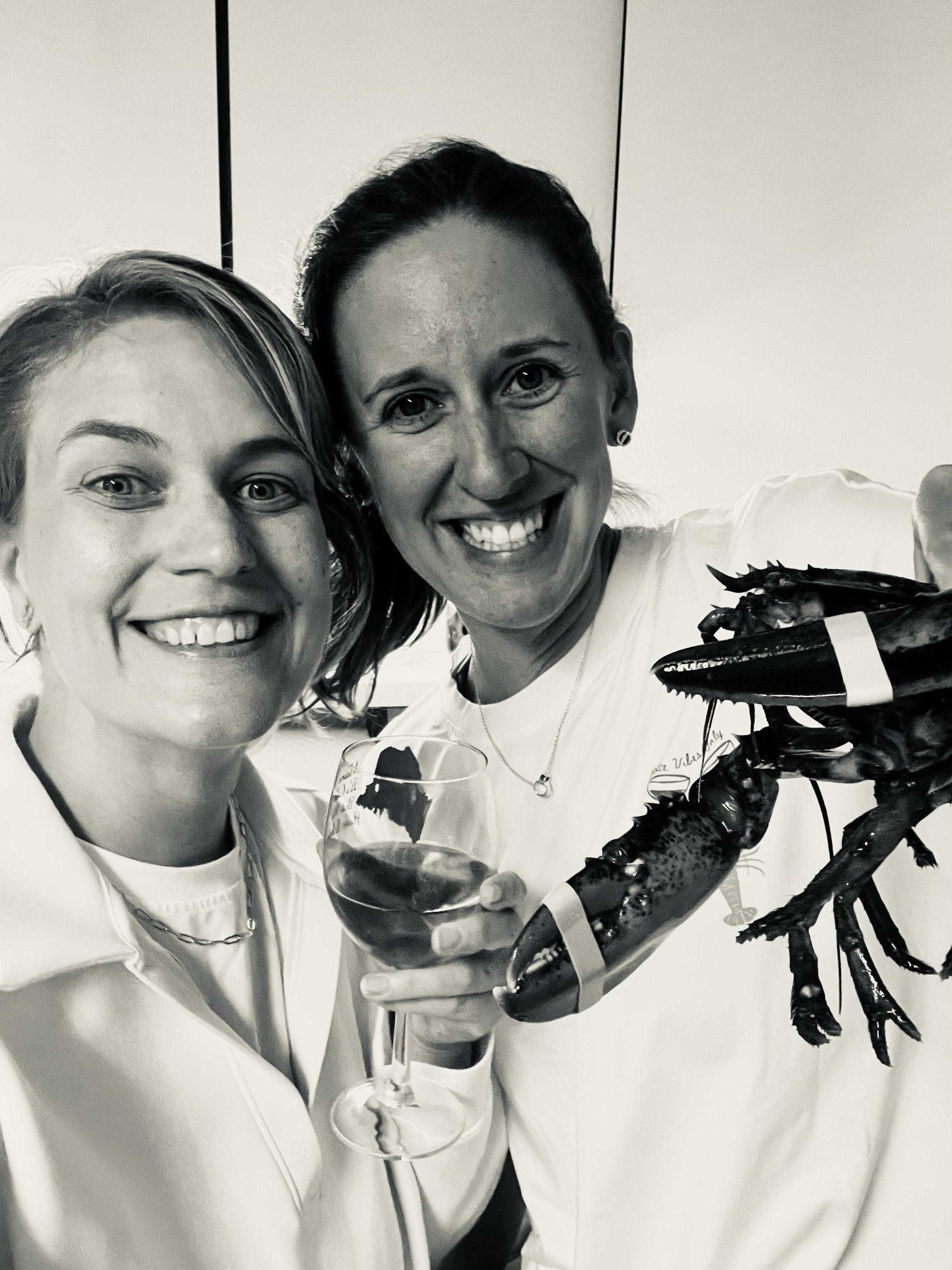 Black and white photo of two smiling female co-founders taking a selfie with one holding a glass of white wine and one holding a lobster.