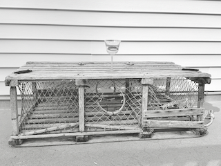 Weathered wooden antique lobster trap with a glass of water on top, set against a white slat wall. Black and white image.