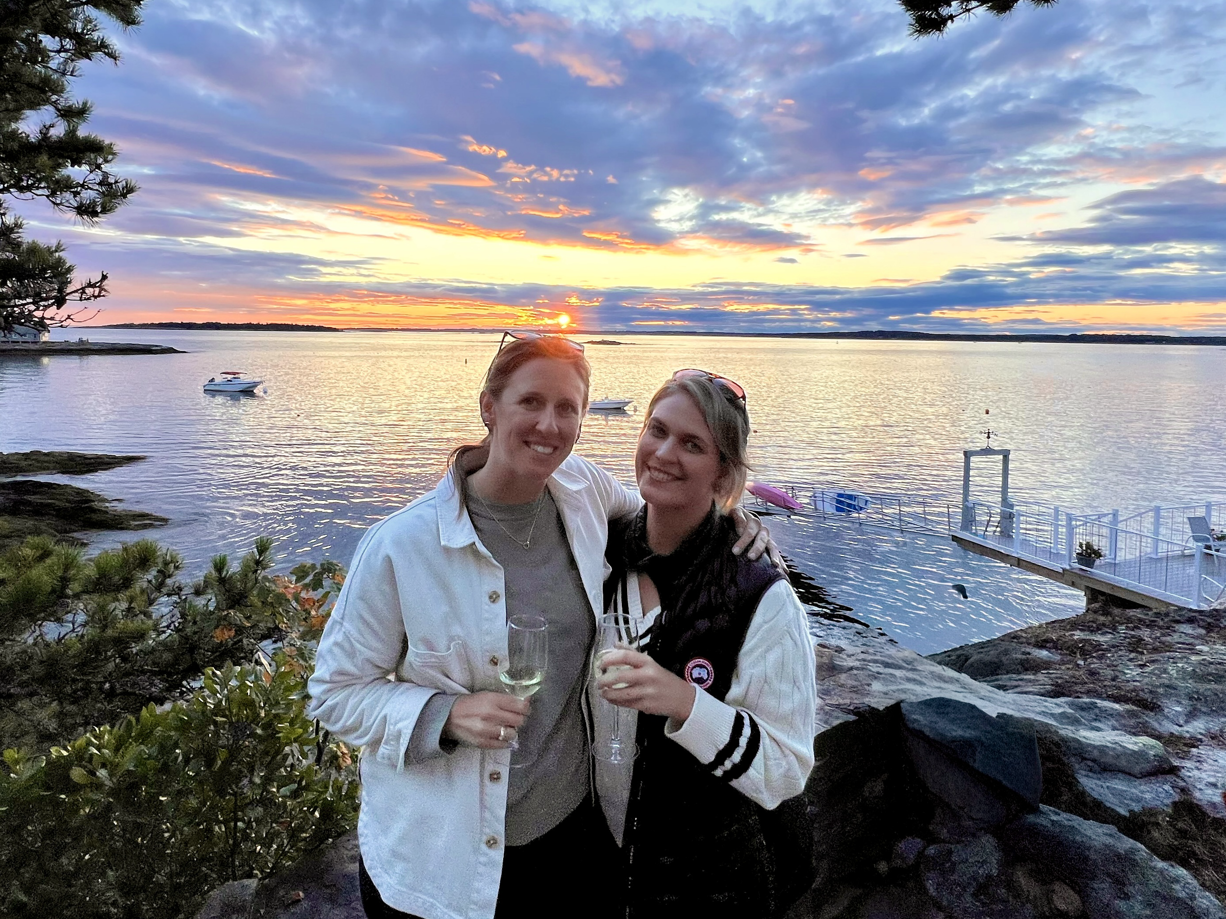 Co-founders standing on the Maine coast at sunset, smiling and holding glasses of wine. The scene includes a calm body of water with boats, a pier, and a colorful sky with clouds and the setting sun.