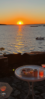 Sunset over the coast of Maine with a boat on water in the background and a table in the foreground holding drinks and snacks.