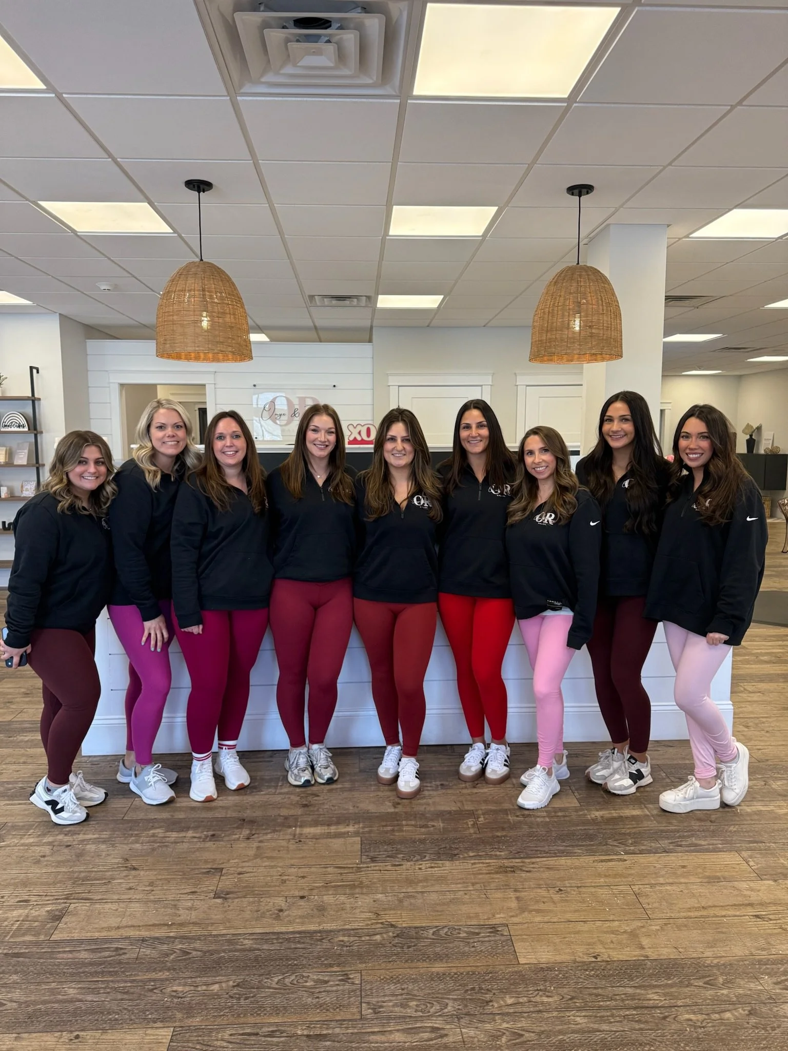 Group of nine women posing together indoors, all wearing black jackets and colorful leggings.