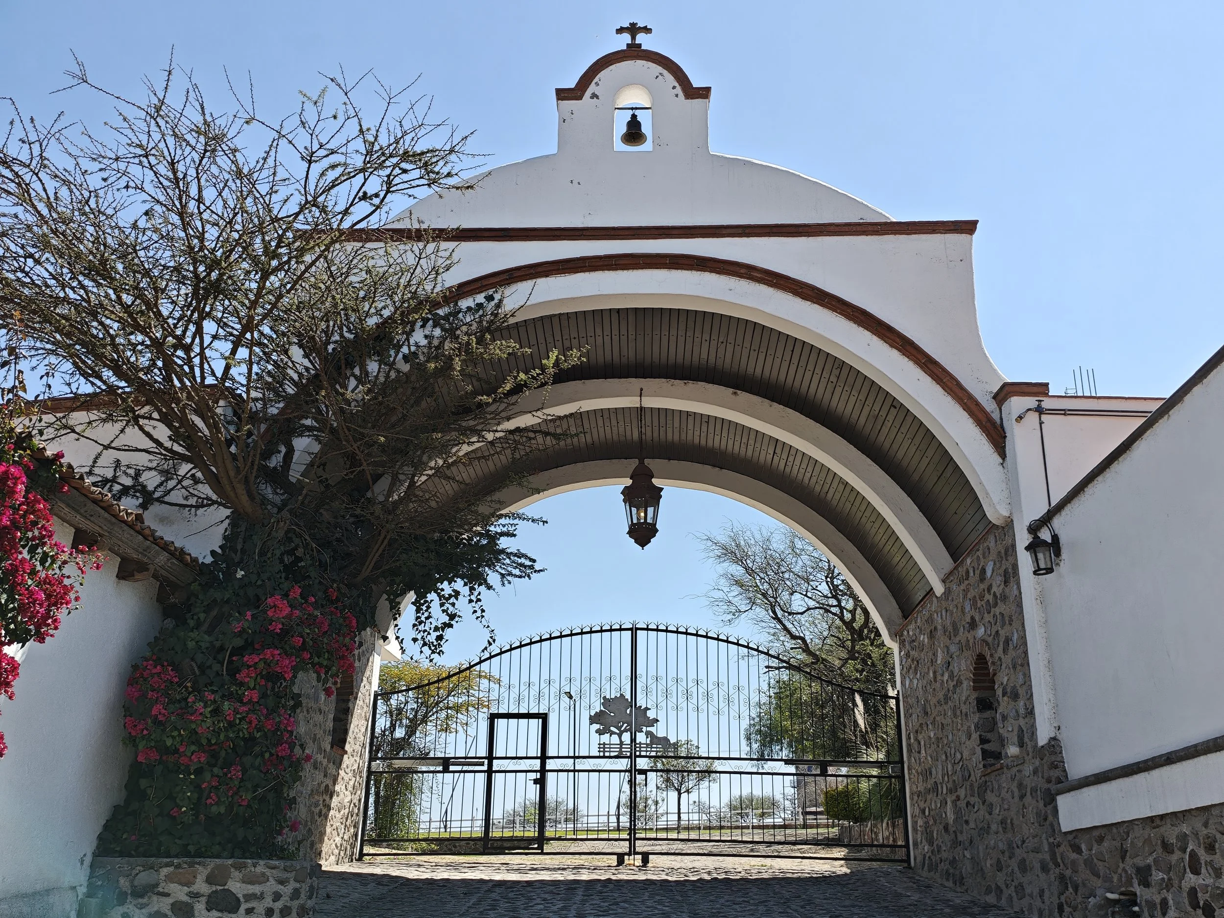 Puerta de entrada a una iglesia con arco y reja, árboles y flores a los lados, y un cielo despejado.