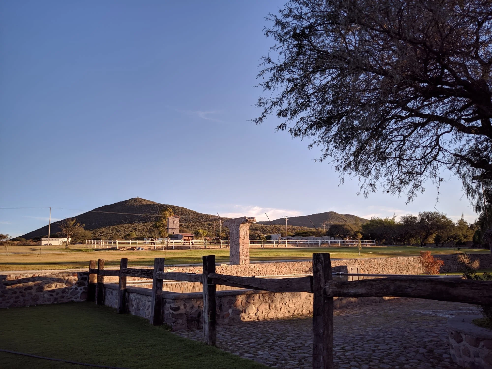 Paisaje rural con montañas en el fondo, árbol grande a la derecha y estructura de piedra en primer plano.