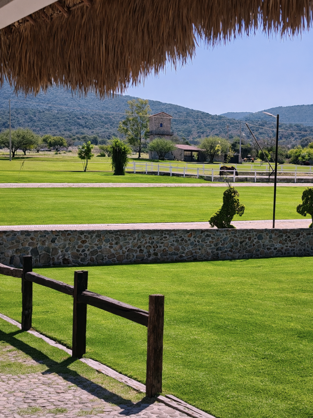 Jardín de césped verde, con un muro de piedra y una cerca de madera, árboles dispersos y un fondo de montañas con un edificio antiguo.