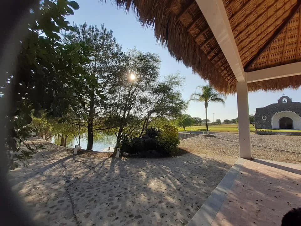 Vista de un jardín con árboles junto a un río, una estructura de techo de paja y una pequeña iglesia al fondo, en un ambiente soleado y tranquilo.