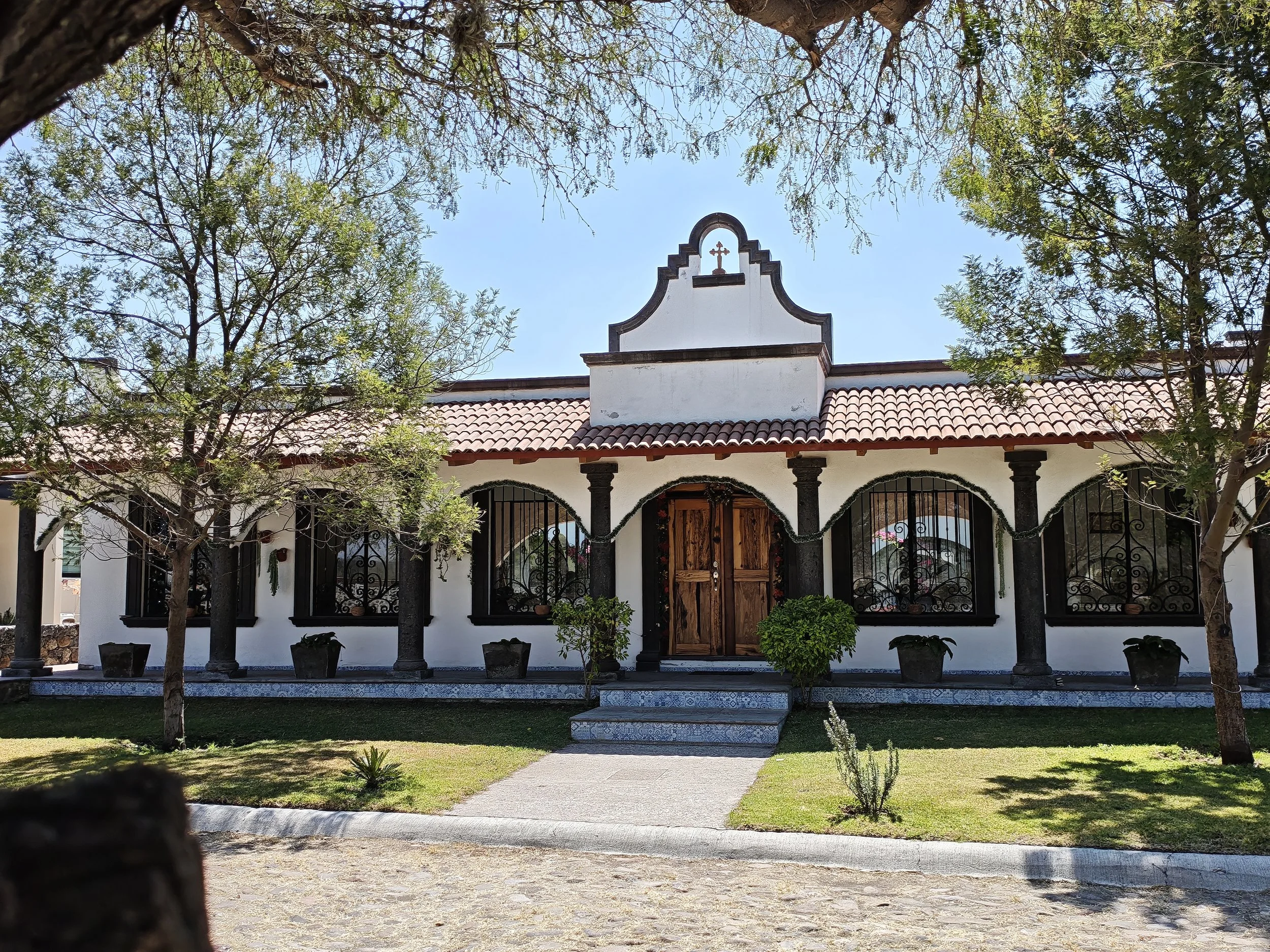 Fachada de una iglesia con puertas de madera, columnas negras y ventanas con rejas decorativas, rodeada de árboles y un césped bien cuidado.