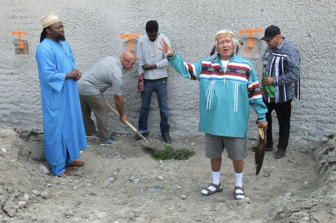 Group of men participating in a groundbreaking ceremony, with one man speaking and others standing around, some in casual and traditional clothing, on a dirt site against a wall with orange spray paint markings.