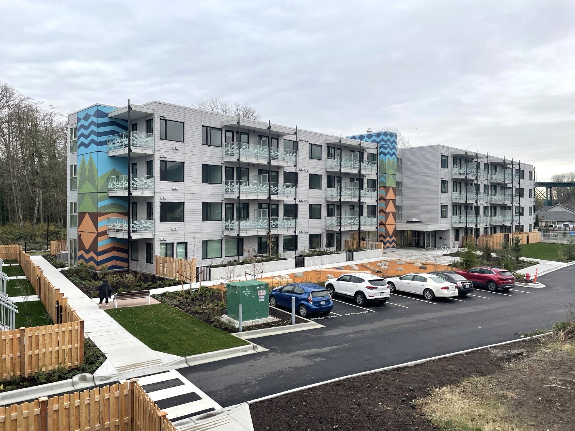 Multi-story residential building with colorful box-like structures protruding from the facade, black-framed windows, glass balconies, a wooden privacy fence, and sidewalk in front, under an overcast sky.