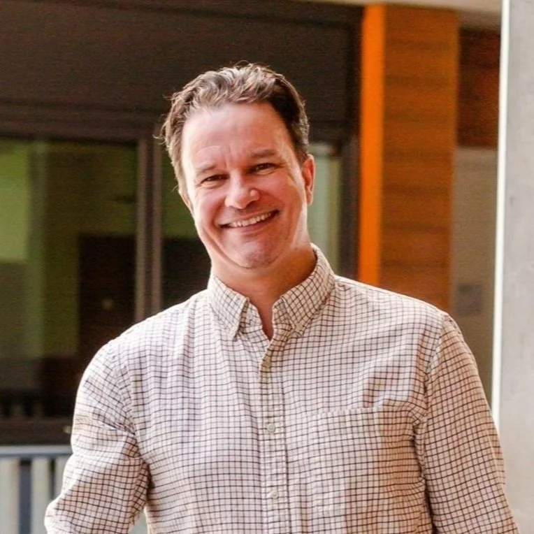 A smiling man with short dark hair wearing a checkered button-down shirt indoors.