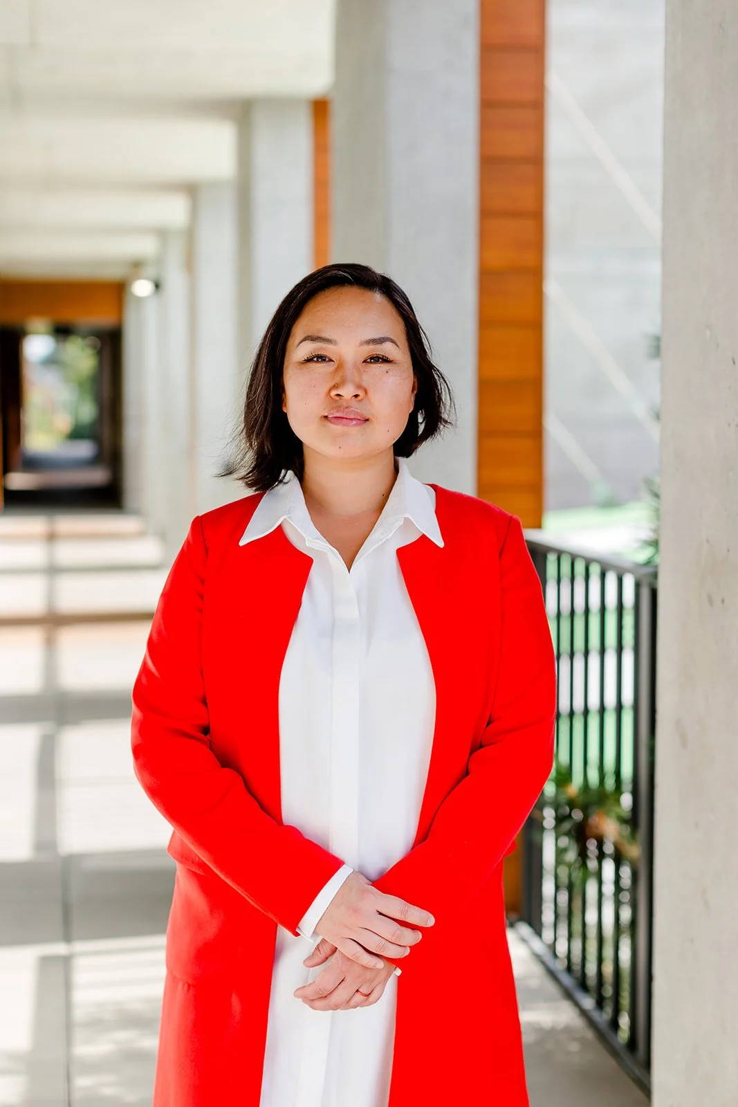 A woman with shoulder-length dark hair, wearing a white blouse and a bright red blazer, standing on a corridor with modern architectural features and natural light in the background.