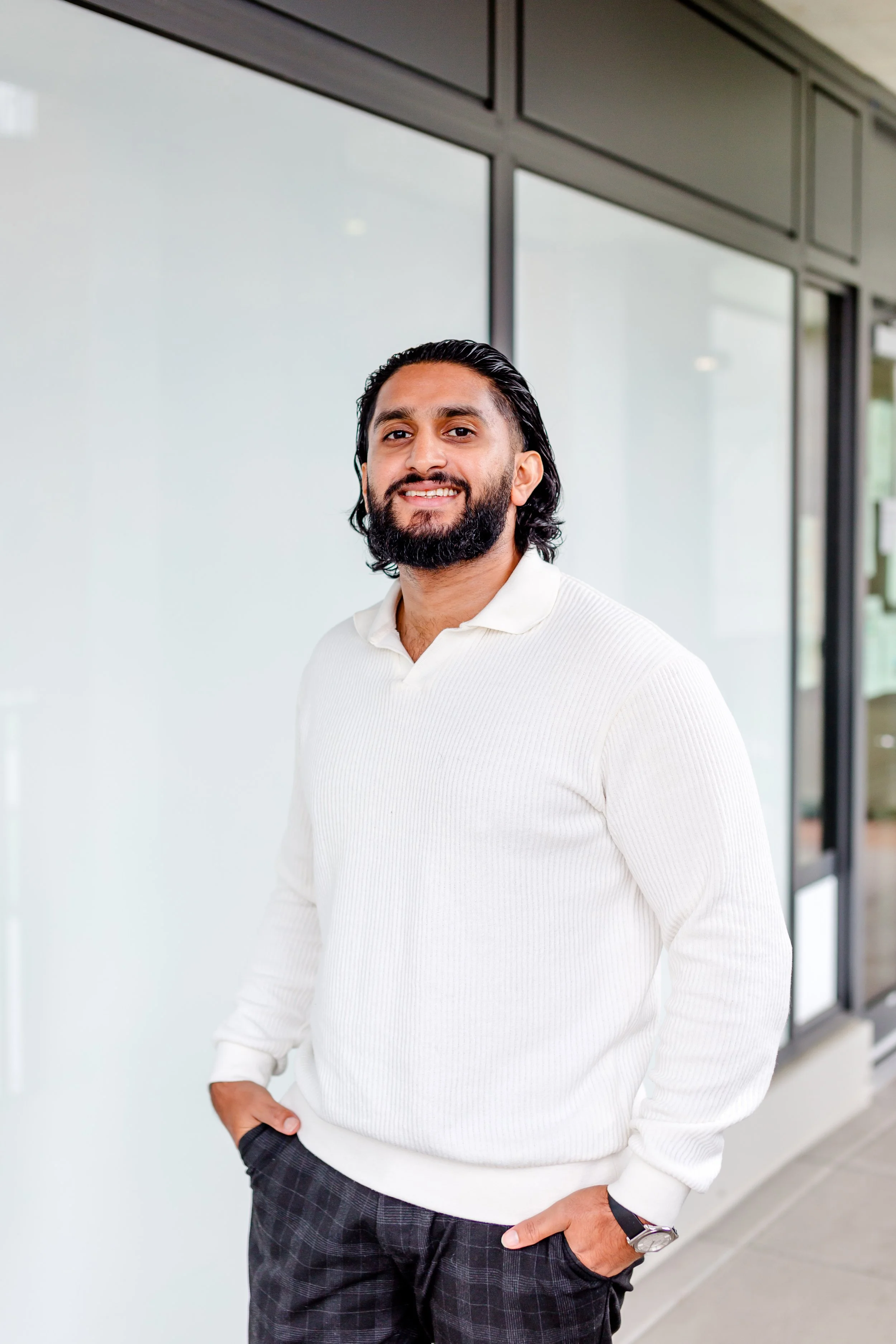 A man with dark hair, a beard, and light brown skin, smiling, standing indoors in front of a glass window with black frames. He's wearing a white ribbed long-sleeve shirt, plaid pants, and a watch on his left wrist. His right hand rests in his pocket, and his left hand is also in his pocket.
