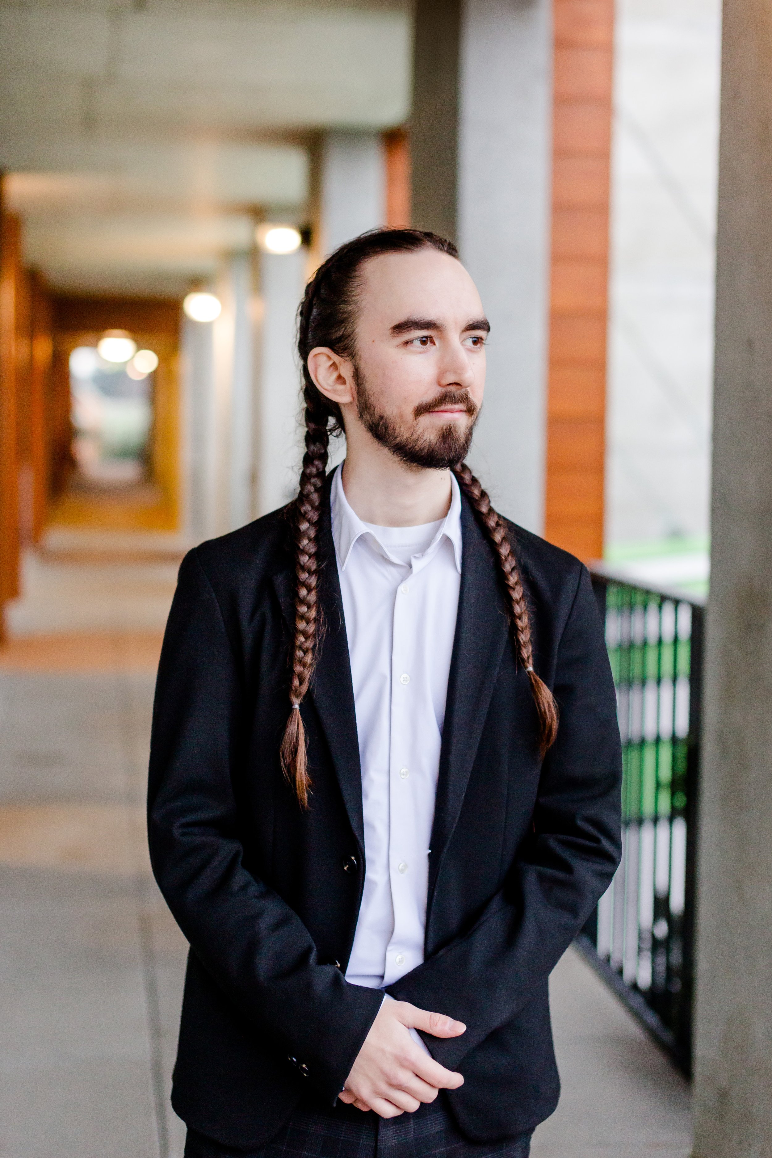 A man with long braided hair and a beard stands outdoors wearing a black blazer over a white shirt, looking to the side with a neutral expression.