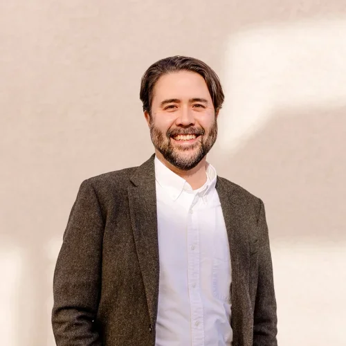A smiling man with dark hair and a beard, wearing a white shirt and a brown blazer, standing against a plain light-colored wall.