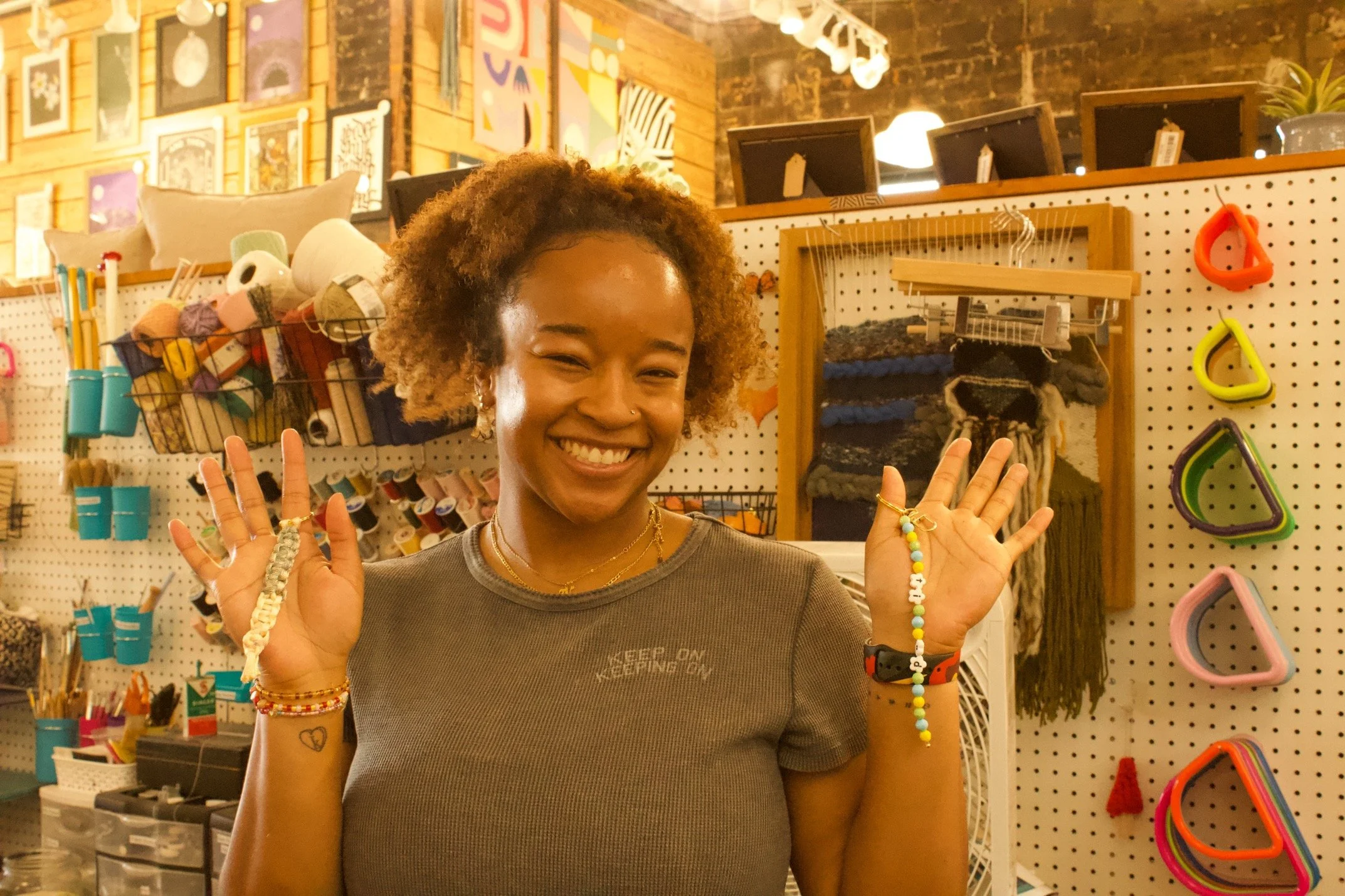 A smiling woman with curly hair showcasing her accessories in a craft store or art supply shop. She is wearing multiple bracelets, rings, and a necklace, and holds colorful beaded jewelry in her hands. The background features shelves with yarn, framed artwork, and various craft supplies.