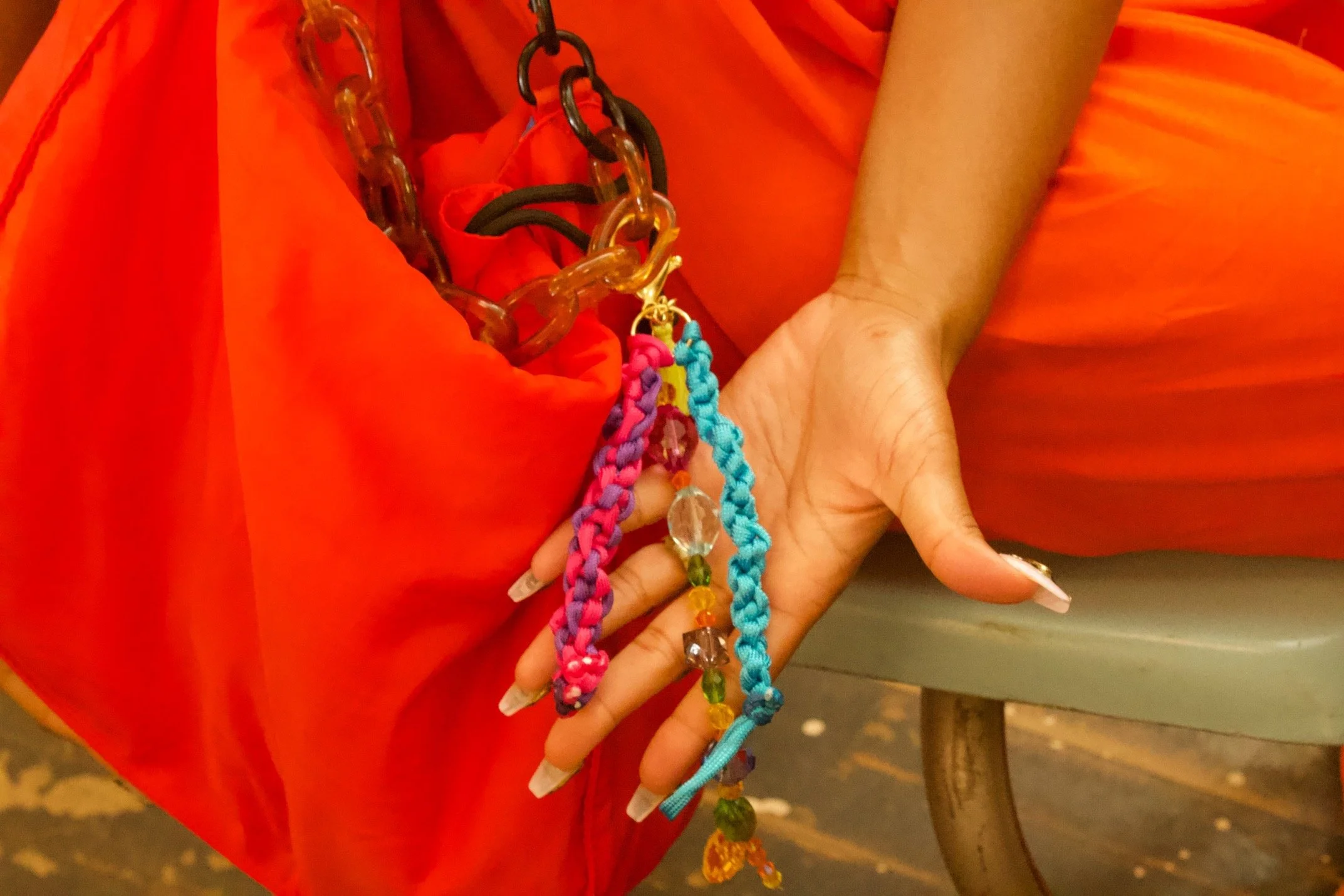 Close-up of a person's hand holding colorful beaded jewelry with a red braided strap, against a background of bright orange fabric.