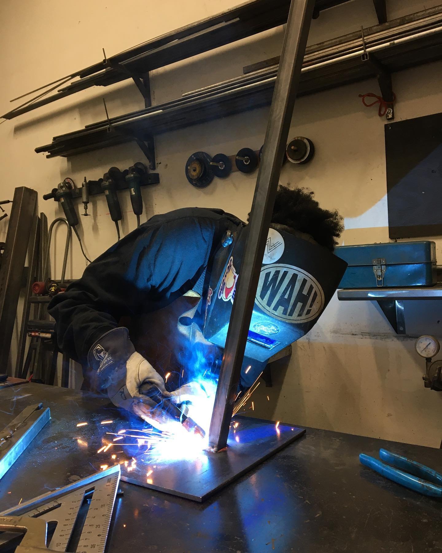 Person welding metal in a workshop, with sparks flying and wearing protective gear including a welding helmet and gloves.