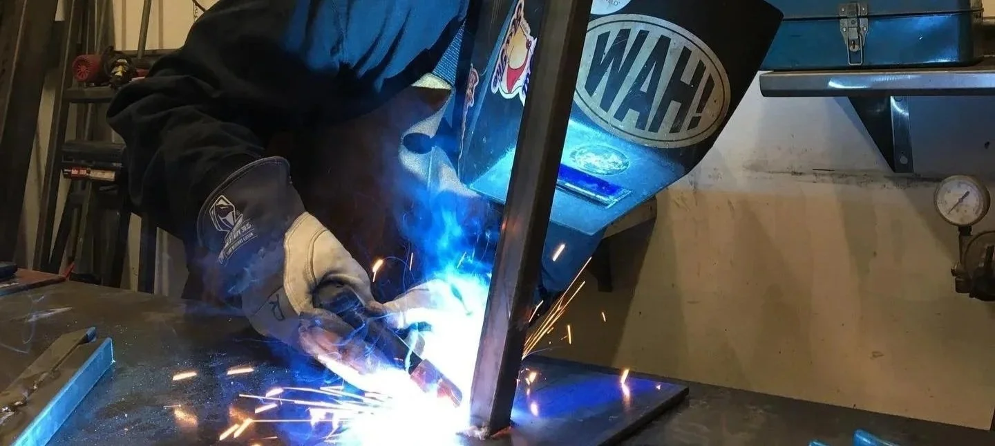 A person welding metal at a workbench in a workshop, with sparks and bright blue light from the welding process.